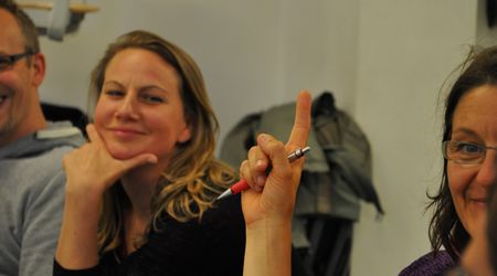 Zwei Frauen sitzen am Tisch, eine grinst in die Kamera, die andere hält ihren Zeigefinger in die Luft