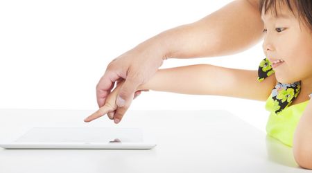 Der Finger einer Kinderhand wird von einer Erwachsenenhand zu einem Tablet geführt